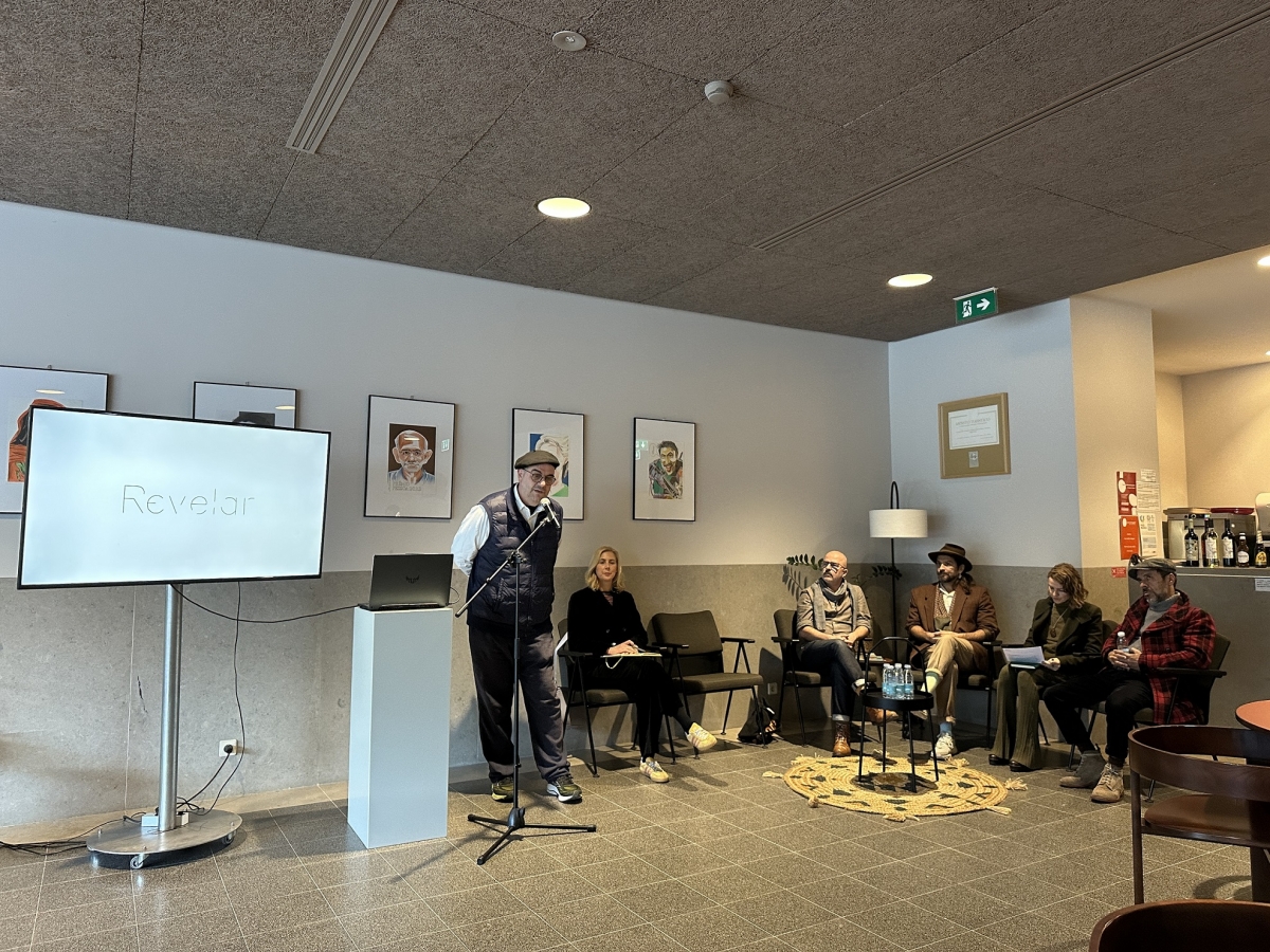 Sessão de apresentação ‘Revelar’ em sala interior: orador ao microfone junto de um ecrã com a palavra ‘Revelar’; cinco pessoas sentadas em pano de fundo.