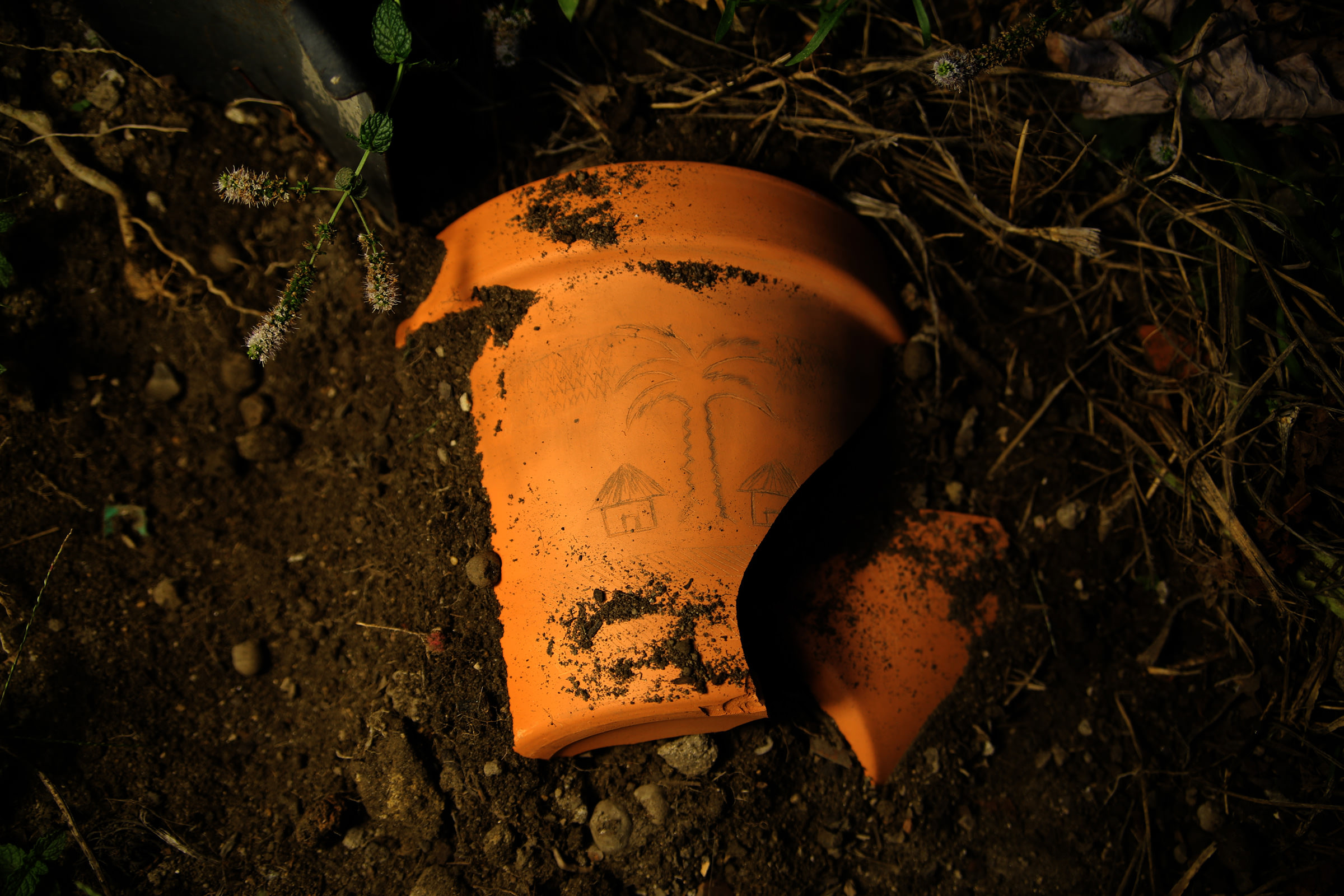 Pedaço de vaso de barro partido no chão, com desenhos de duas casas e uma palmeira. Fotografia noturna com luz dramática e texturas realçadas.